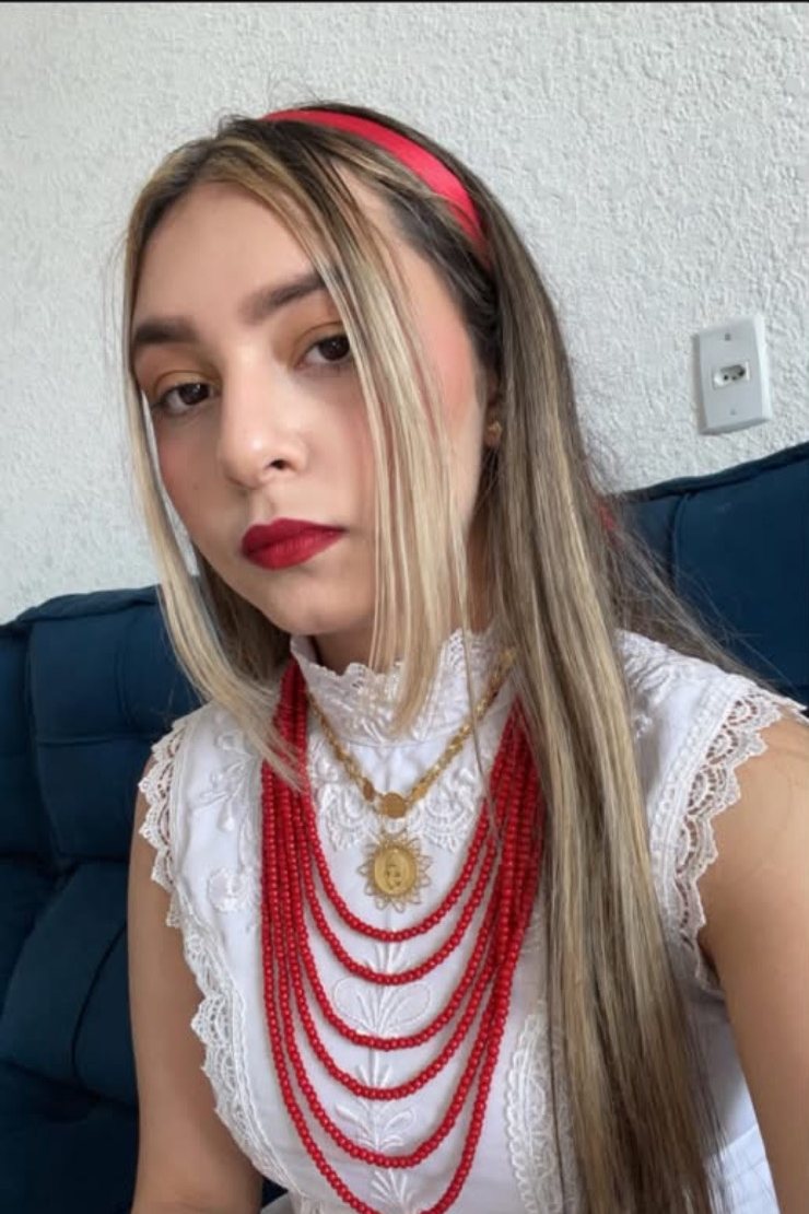 Mãe Lavínia de Rosa Caveira de vestido branco de renda, fita vermelha no cabelo e guias vermelhas no pescoço